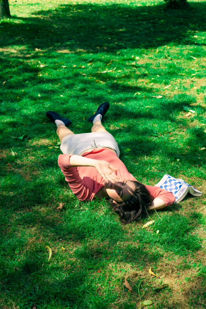 Een jonge vrouw ligt in het gras en houd haar hand voor haar gezicht als bescherming tegen de zon.